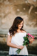 Beautiful young woman with spring flowers bouquet at city street. Happy girl smiling and holding pink roses flowers outdoors. Spring portrait of pretty female.