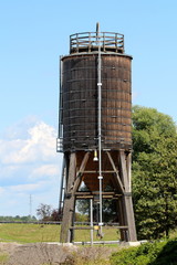 Old wooden storage silo with metal support frame and narrow metal pipe attached to one side at local construction site surrounded with grass and tall trees