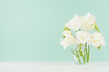 Fresh white freesia flowers in cute transparent glass on soft light green mint menthe wall and white wood table, copy space. - obrazy, fototapety, plakaty