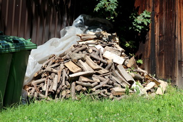Pile of freshly cut firewood stacked on top of transparent nylon and uncut grass in corner of suburban family house backyard surrounded with tall wooden and metal fence