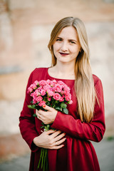 Portrait of young stylish beautiful girl in a red dress with bouquet of flowers stand on the street, smiling enjoy her weekends. Summer, sunny, trendy, lifestyle.