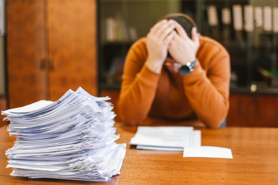 A businessman grabs his head from a pile of papers at work. The concept of depression from difficulties and possible bankruptcy of an enterprise.