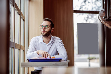 Young thoughtful man elegantly dressed sitting in fast food restaurant, having lunch break and...