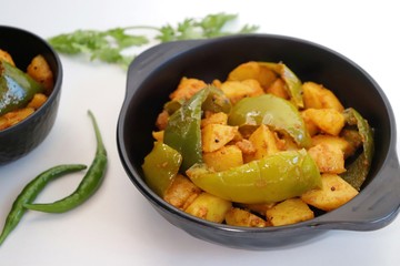 Dry capsicum and potatoes curry. also known as Aloo shimla mirchi Masala. served in black bowl with chilies and coriander over white background with copy space.