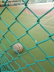 An old, used, dirty basketball behind a wire fence; on a basketball field; inside a school. A motorcycle parking space is visible in the background.