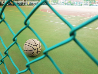 An old, used, dirty basketball behind a wire fence; on a basketball field; inside a school. A motorcycle parking space is visible in the background.