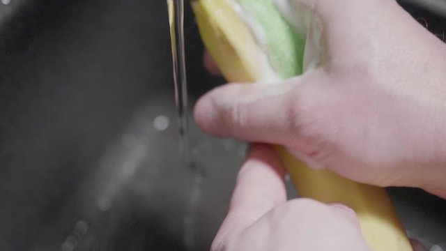 Man washes banana with washing supplier and sponge. Foam is on hands and banana. Black sink is on background. Close up and slow motion view.