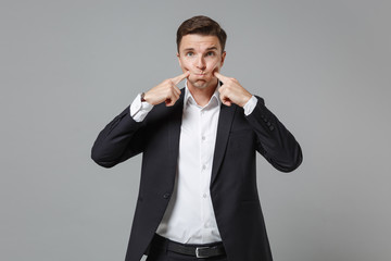 Young business man in classic black suit shirt posing isolated on grey background in studio. Achievement career wealth business concept. Mock up copy space. Pointing index fingers on blowing cheeks.