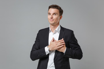 Pleasant young business man in classic black suit shirt posing isolated on grey wall background. Achievement career wealth business concept. Mock up copy space. Holding hands crossed on chest, heart.