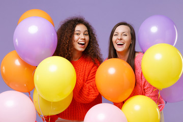 Cheerful two european african american women friends in knitted sweaters isolated on violet purple background. Birthday holiday party, people emotions concept. Celebrating hold colorful air balloons.