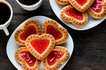 Heart-shaped cookies with strawberry jam on a white plate and two white cups of coffee on a brown wooden background. Love concept. View from above.