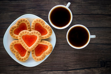 Heart-shaped cookies with strawberry jam on a white plate and two white cups of coffee on a brown wooden background. Love concept. Top view.