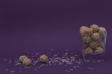 Natural energy balls in coconut flakes in a transparent glass cup. Three balls and coconut flakes lie next to the glass. Horizontal view. Side view. Copy space. Dark background.