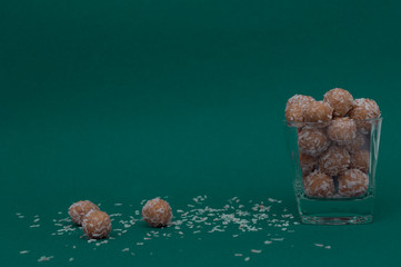 Dietary healthy snack in coconut flakes in a transparent glass cup. Three energy balls and coconut flakes lie next to the glass. Horizontal view. Side view. Copy space. Dark background.