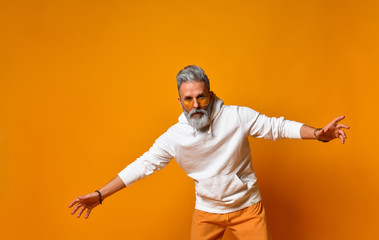 Elderly male in white hoodie, pants and sunglasses, bracelets. He spread his arms and posing against orange background. Close up