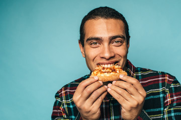Young latin hispanic man stand on right side of picture and smile. Hold one slice of pizza in hands and bite eat. Eating fatning unhealthy delicious meal. Isolated over blue background.