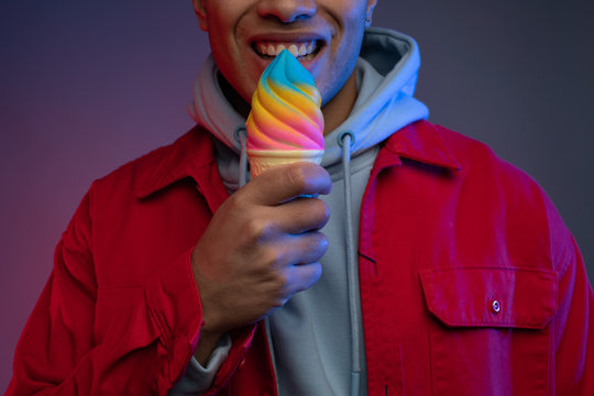 Dark picture of cheerful young arabian man hold colorful ice cream and smile. Getting ready to eat it. Guy wear stylish grey hoodie and red jacket. Isolated over colorful background. Cut view.