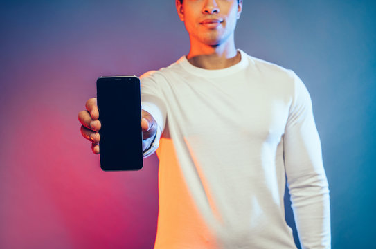 Cut view of young arabian man hold black phone in hand and show it to camera. Strong powerful man wear white sweater. Modern technologies. Isolated over colorful background.