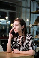 Stylish young beautiful smiling girl in a striped shirt and black dress is sitting at a table in a cafe and talking on the phone. 