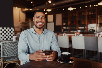 Happy african young man sitting in cafe using mobile phone looking at camera