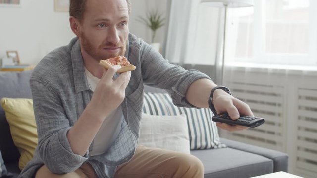 Tilt up of ginger bearded man wearing comfortable clothes sitting on sofa, eating pizza while watching football match on TV