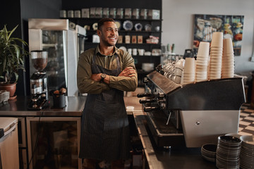 Thoughtful afro-american small coffee shop owner standing behind counter wearing apron with crossed...