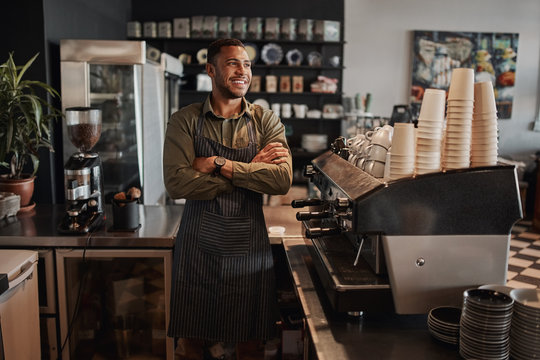 Thoughtful afro-american small coffee shop owner standing behind counter wearing apron with crossed arms looking away