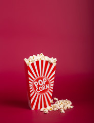 Popcorn in striped bucket on red background. Hot corn scattered from the paper box, copy space. Fast food and movie snack.