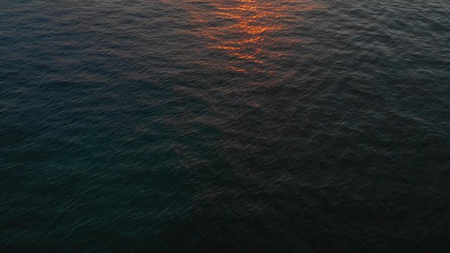A drone skims the ocean off the coast of Oahu showing the colors weaved in between the ripples of the wind kissed ocean. Pans up to show a sunset.