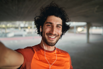 Portrait of a smiling fit young man with earphones in his ears taking selfie outdoors - pov shot of...