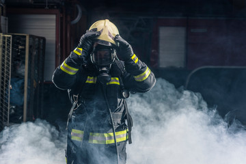 Fireman standing confident holding helmet and wearing firefighter turnouts. Portrait of a fireman with dark background with smoke and blue light.