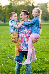 Fototapeta premium Family portrait of three. Happy father hugs his brunet small son and blond daughter outdoors in city park on nature. On background sunset. Happy loving family concept. all smiling