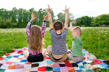 Back view of children spending time outside, boys and girl sitting on plaid raise their hands up and show gestures. Happy childhood concept. Cheerful children resting on backyard