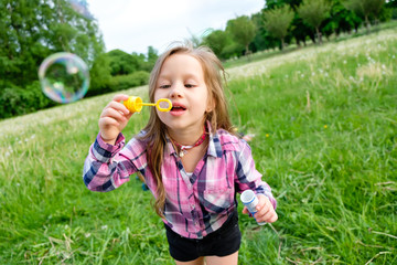 Cute little girl in shirt and shorts is blowing soap bubbles in park summer day on camera