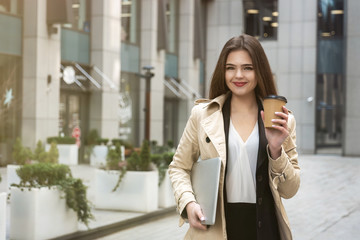 Fototapeta premium Young business lady holding laptop under arm walking down the street near office center with cup of coffee looking busy, eating on the go concept