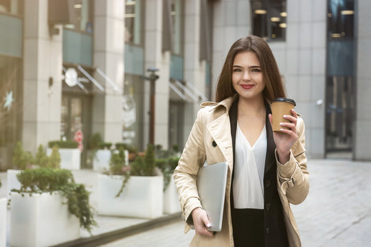 Young Business Lady Holding Laptop Under Arm Walking Down The Street Near Office Center With Cup Of Coffee Looking Busy, Eating On The Go Concept