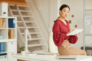 Portrait of elegant Asian businesswoman sitting on desk and looking at camera while working in...