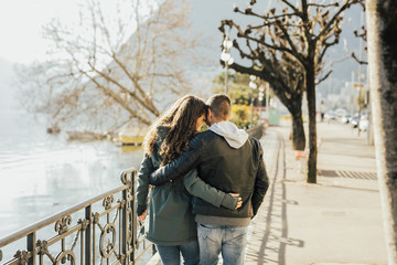 Back view of couple hugging and walking on the shore of lake Lugano, Switzerland. Lovers in the sunny day.
