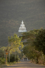 long street road heading to an entrance of thai temple which big white leg crossed budda statue is on a green mountain