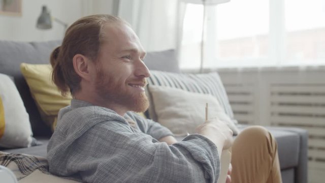 Close up of Caucasian man with red hair wearing comfortable clothes sitting in living room, eating Asian food from box while watching television