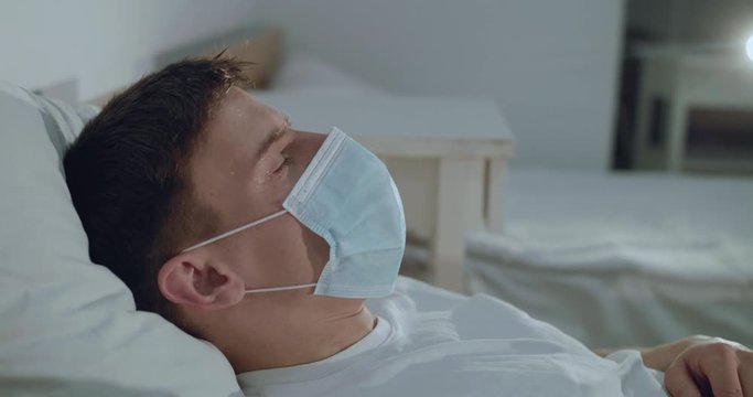 Portrait of ill, dying from virus man in medical mask lying and coughing on bed