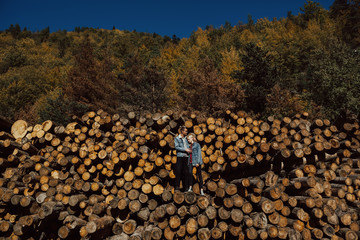 Couple enjoying their holiday in a mountain. Boy and girl standing on the stack of firewood in the autumn forest. A walk of a couple in love in a beautiful colorful forest. 