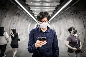 Asian man wearing surgical face mask using smartphone in subway tunnel with crowded people walking. Wuhan coronavirus (COVID-19) outbreak prevention in public area. Health care and medical concept