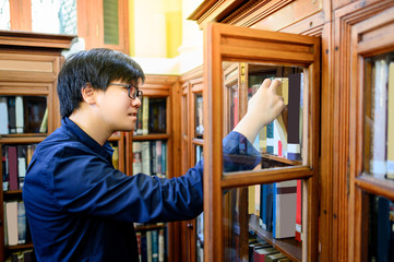 Asian man university student hand choosing and picking vintage book from old wooden bookshelf in college library. Antique textbook resources for education research. History and literature learning