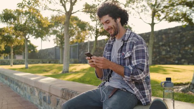 Portrait of a handsome smiling young man sitting in the park with his water bottle and earphones in his ears texting messages on smartphone