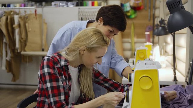 Side view tilt down shot of young seamstress learning to use sewing machine helped by cheerful middle aged colleague in sewing studio