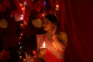 Obraz na płótnie Canvas An young and beautiful Indian Bengali woman in Indian traditional dress is sitting while holding Diwali diya/lamp in her hand in front of colorful bokeh lights. Indian lifestyle and Diwali celebration