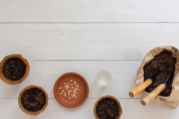 Eco friendly pots for seedlings and small bag with ground and a garden shovel and rakes on a white wooden background, planting cucumber seeds, top view