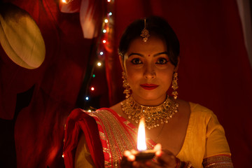 Obraz na płótnie Canvas An young and beautiful Indian Bengali woman in Indian traditional dress is sitting while holding Diwali diya/lamp in her hand in front of colorful bokeh lights. Indian lifestyle and Diwali celebration