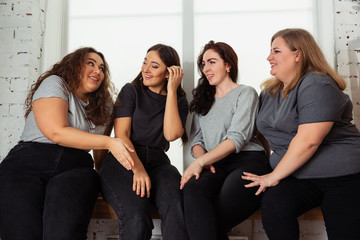 Young caucasian women in casual clothes having fun together. Friends sitting near by window and laughting, spending time together. Bodypositive, nutrition, feminism, loving themself, beauty concept.